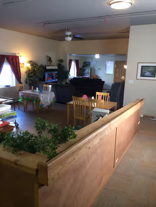 Interior view of a senior living facility common area with wooden furniture including tables and chairs, a couch, plants, and a television. The room has beige walls, red curtains on the windows, and a ceiling fan with a light fixture.