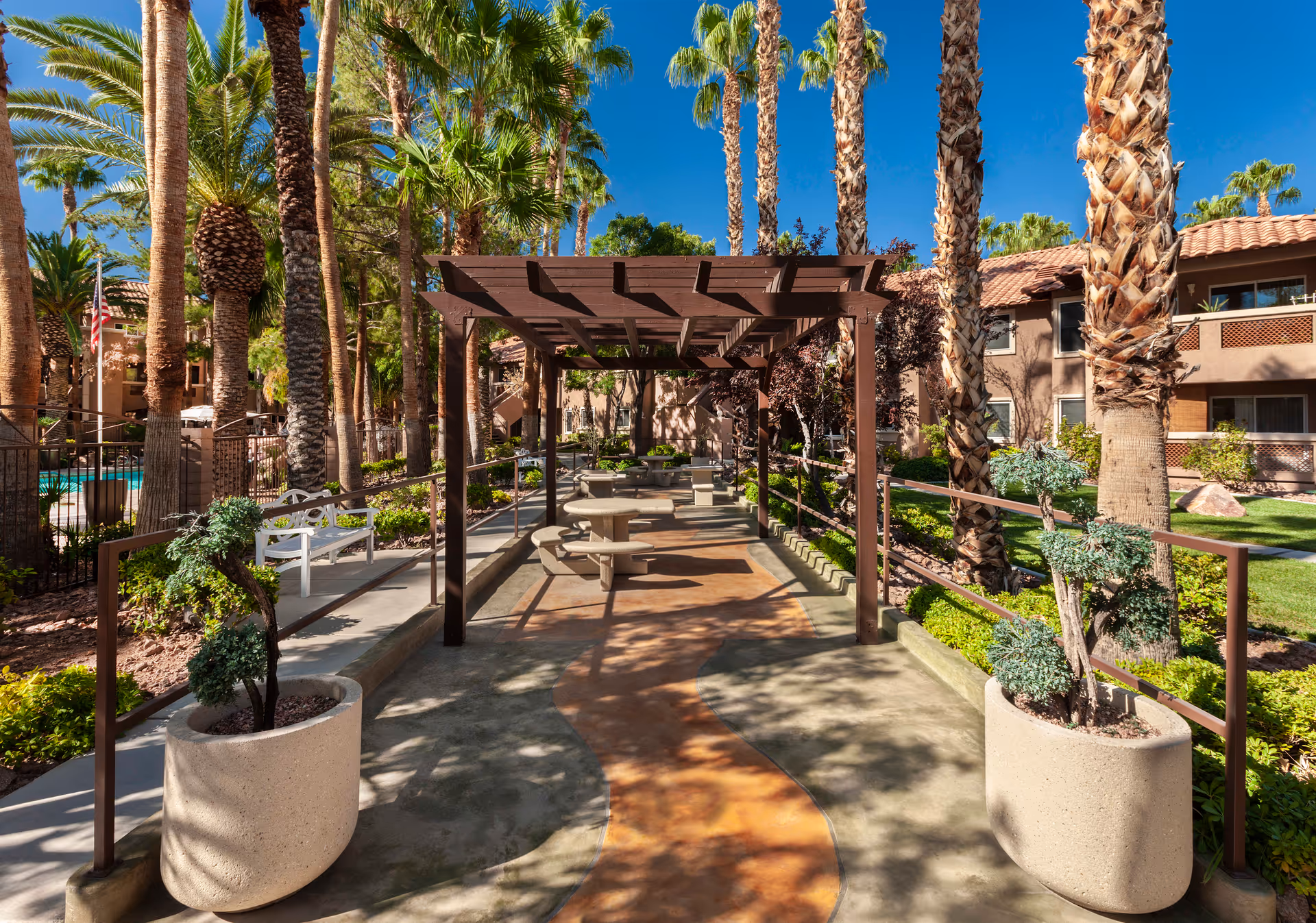 Outdoor seating area at Destinations Spring Valley featuring a shaded pergola with round concrete tables and benches, surrounded by palm trees, landscaped greenery, and residential buildings under a clear blue sky.