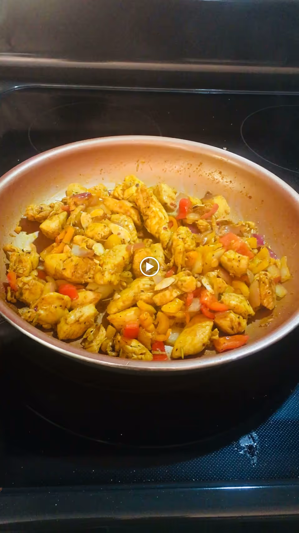 A frying pan on a stovetop with cooked pieces of chicken mixed with chopped vegetables including red and yellow bell peppers and onions.