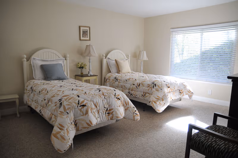 A bright bedroom with two twin beds featuring white headboards and floral-patterned bedding. Each bed has a pillow and a cushion. Between the beds is a small nightstand with a lamp and a small flower arrangement. A window with blinds lets in natural light, and there is a chair with a patterned cushion in the foreground.