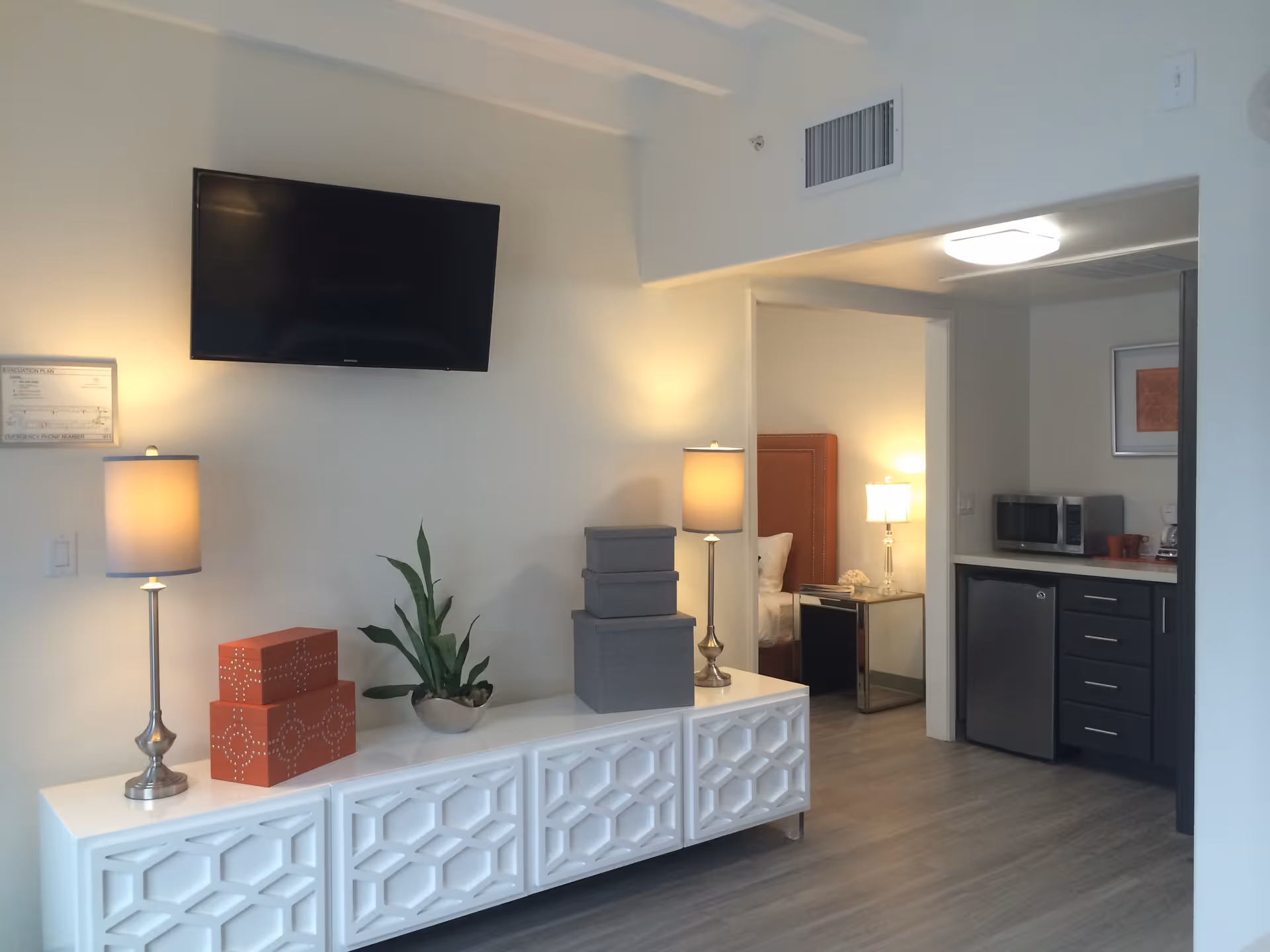 Interior view of a senior living facility room showing a white cabinet with decorative boxes and a plant on top, two table lamps, a wall-mounted flat-screen TV, and a small kitchenette area with a microwave, mini fridge, and coffee maker. In the background, a bedroom with a bed, nightstand, and lamp is visible.