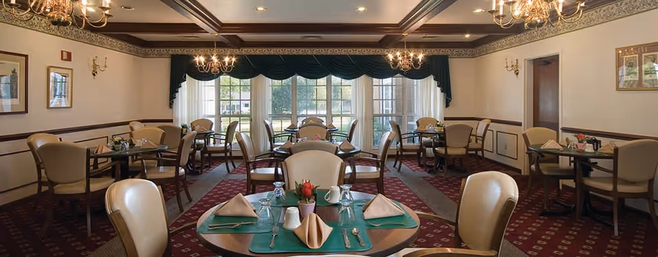 A well-lit dining room with multiple round tables set with green placemats, beige napkins, glassware, and silverware. The room features beige upholstered chairs, chandeliers hanging from a coffered ceiling, wall sconces, framed artwork, and large windows with dark green valances allowing natural light to enter.