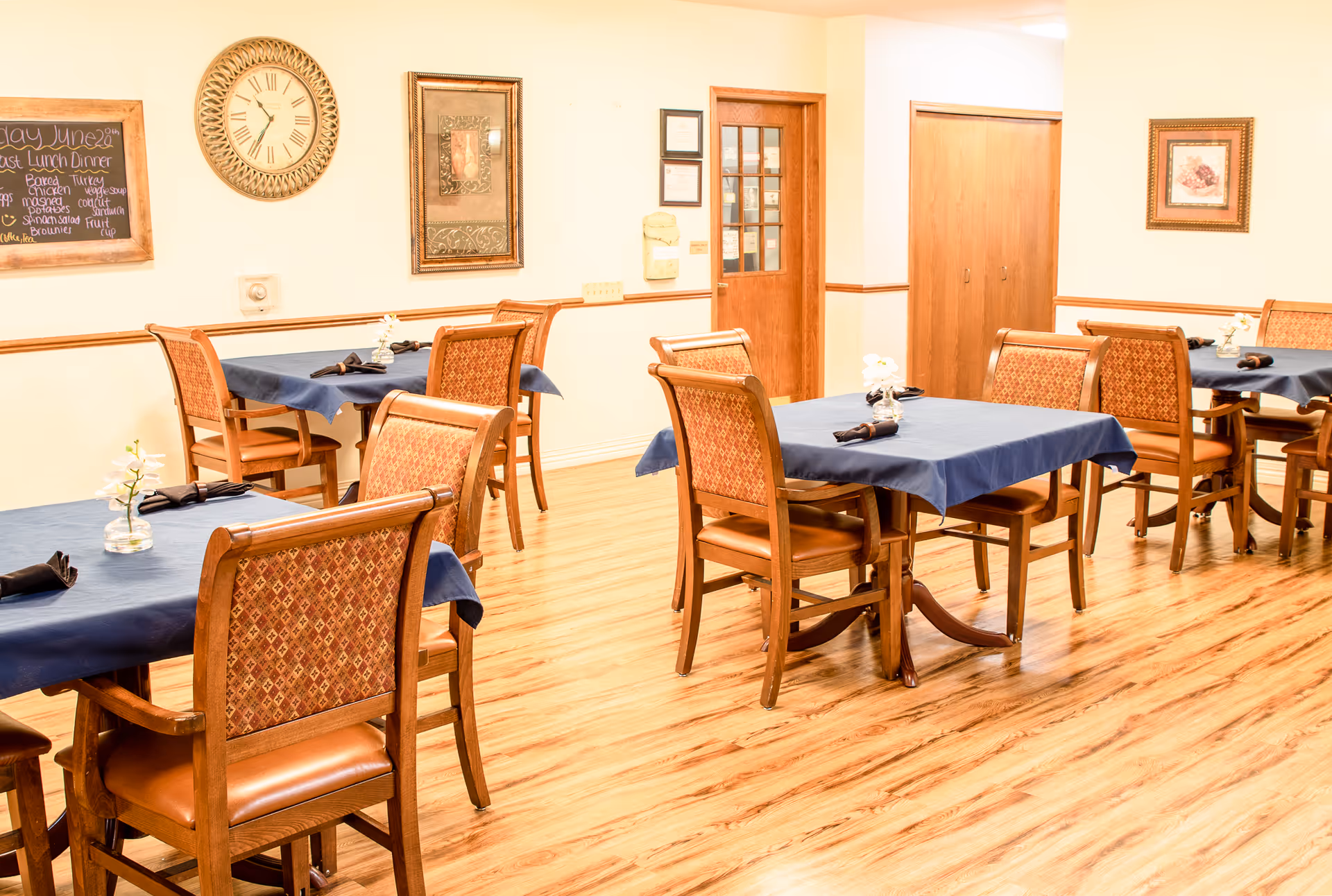 A dining room with wooden floors and several tables covered with blue tablecloths. Each table is surrounded by wooden chairs with patterned upholstery. The walls are decorated with framed pictures, a clock, and a chalkboard menu listing lunch and dinner options. The room has a warm, inviting atmosphere.