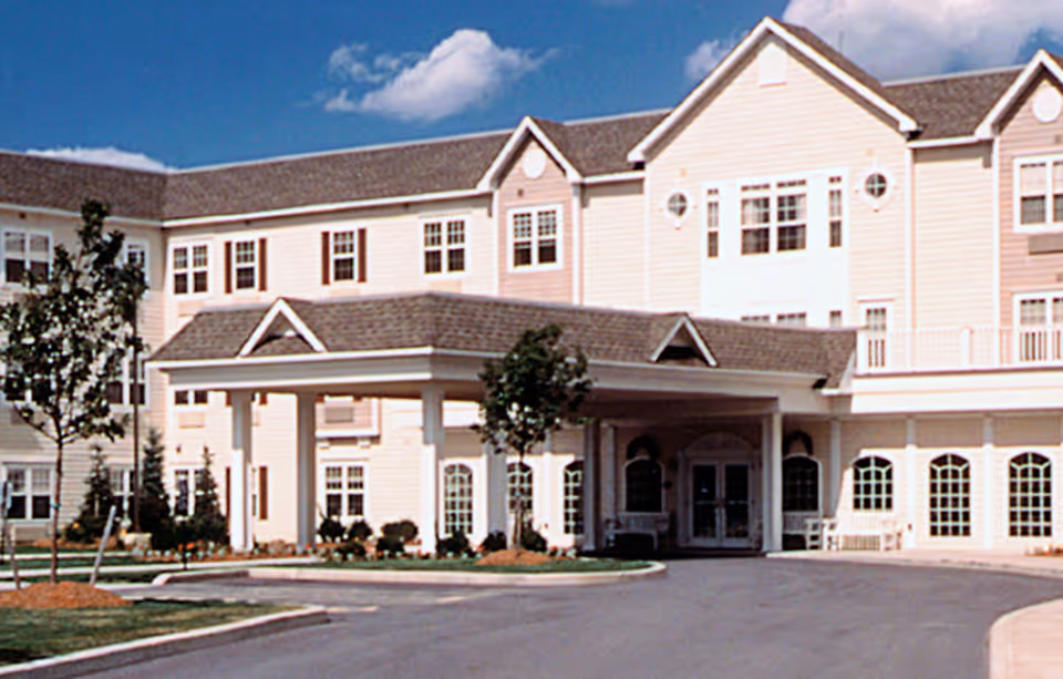Front entrance of a multi-story beige assisted living building with a covered porte-cochère and paved driveway.