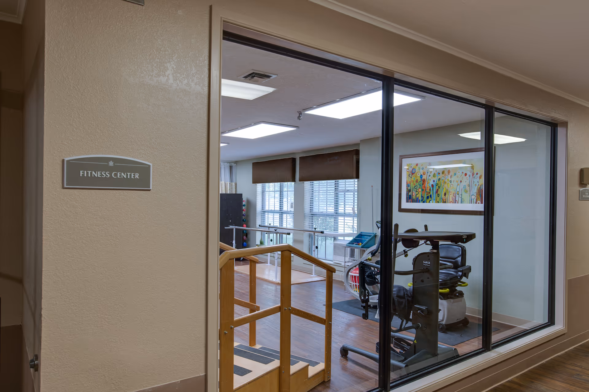 View through a glass wall into a fitness center with exercise equipment, wooden handrails and large windows and a 'FITNESS CENTER' sign on the wall.