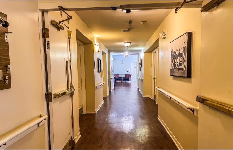 A well-lit hallway in a senior living facility with wooden flooring, beige walls, handrails on both sides, and a few framed pictures on the walls. At the end of the hallway, there are two chairs and a small table near a window.