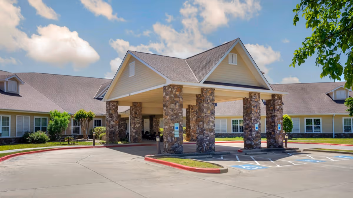 Exterior view of Brookdale Chenal Heights senior living facility showing a covered entrance supported by stone pillars, with a driveway and handicap parking spaces in front, surrounded by a well-maintained lawn and trees under a partly cloudy sky.