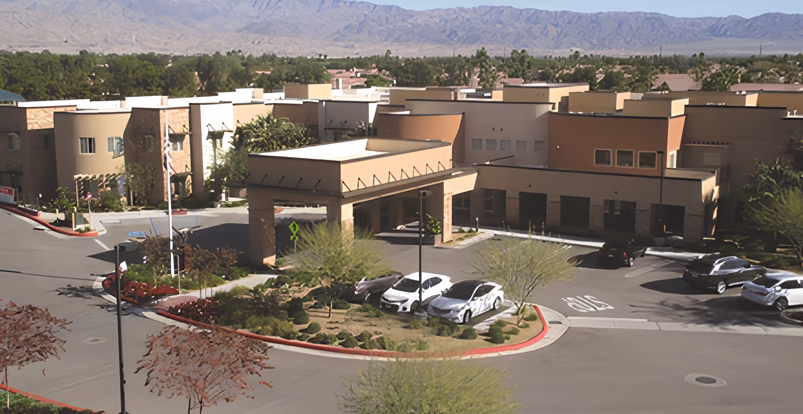 Exterior view of The Palms At La Quinta Gracious Retirement Living facility showing a modern building with a covered entrance, parking spaces with several cars, landscaped greenery, and mountains in the background under a clear sky.