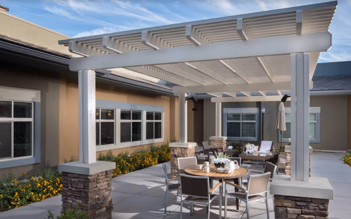 Outdoor patio area at Avenir Memory Care Westside featuring a white pergola with stone pillars, round wooden table with chairs, and additional seating with cushions and a closed umbrella. The patio is surrounded by a building with windows and flower beds with yellow flowers.