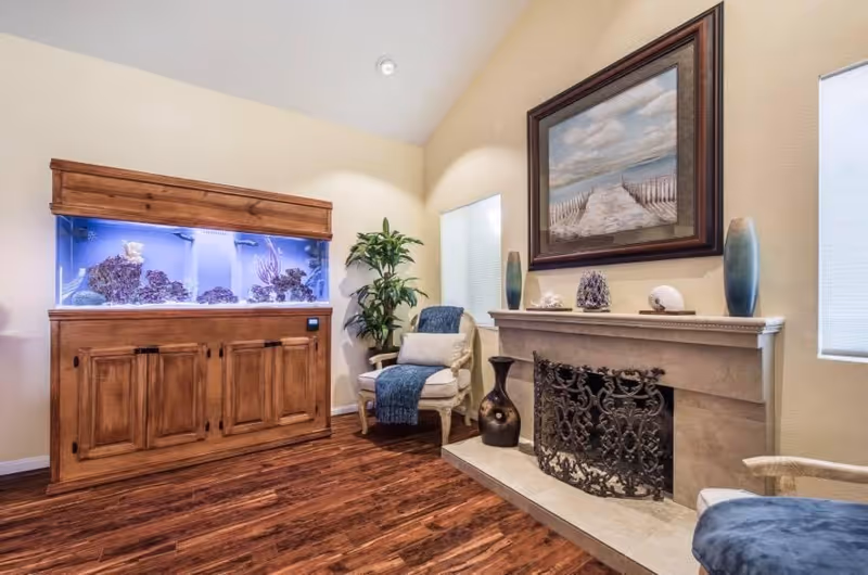 A cozy living room area featuring a large built-in aquarium with coral decorations, a comfortable armchair draped with a blue throw, a fireplace with an ornate metal screen, decorative vases, and a framed painting of a beach scene on the wall.