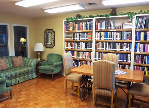 A cozy room with wooden parquet flooring featuring a green patterned sofa, a green armchair, a wooden table with four striped upholstered chairs, and a large bookshelf filled with books against a yellow wall. There is a lamp on a side table and a decorative mirror on the wall.