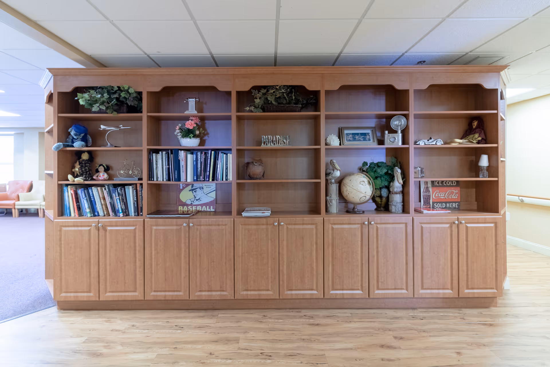 A large wooden bookshelf with multiple compartments containing books, decorative items such as a globe, small statues, plants, a model airplane, and framed pictures. The bookshelf is situated in a well-lit room with light-colored walls and wooden flooring. In the background, there is a glimpse of a seating area with chairs.