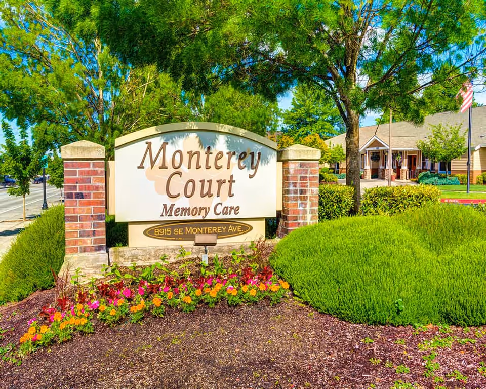 Entrance sign reading "Monterey Court Memory Care" surrounded by flowers and shrubs with the facility building visible behind it.