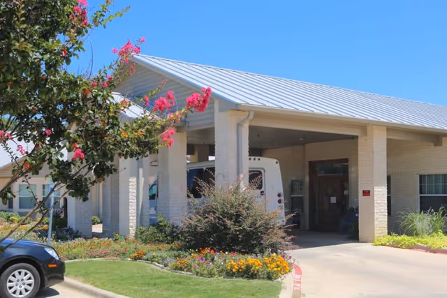 Entrance of a single-story rehabilitation and healthcare center building with a covered drop-off area, surrounded by landscaped flower beds and greenery under a clear blue sky.