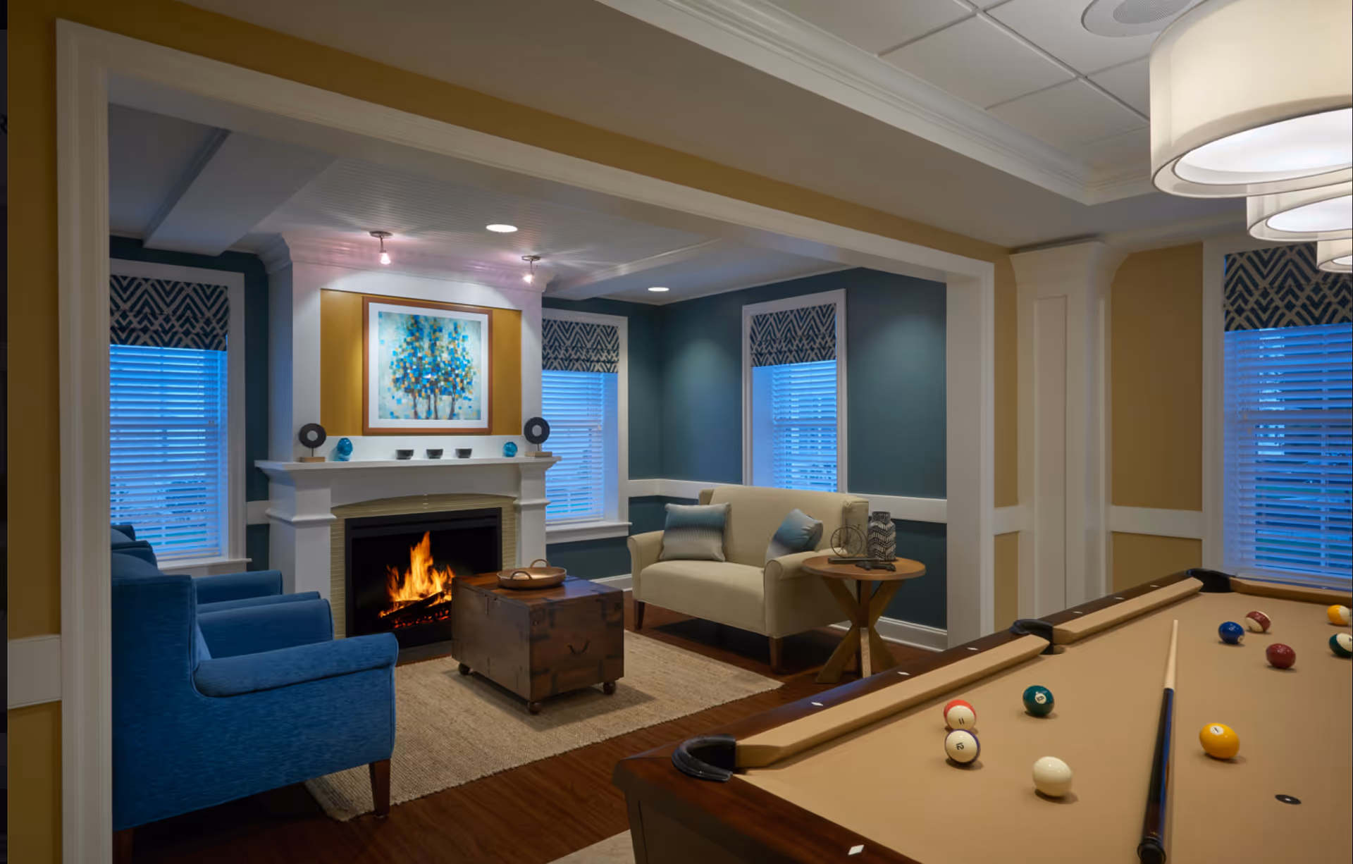 A cozy living room area with a lit fireplace, a beige loveseat with blue pillows, two blue armchairs, a wooden chest coffee table, and a pool table in the foreground. The walls are painted blue and beige with white trim, and there are three windows with patterned blinds. A colorful abstract painting hangs above the fireplace.