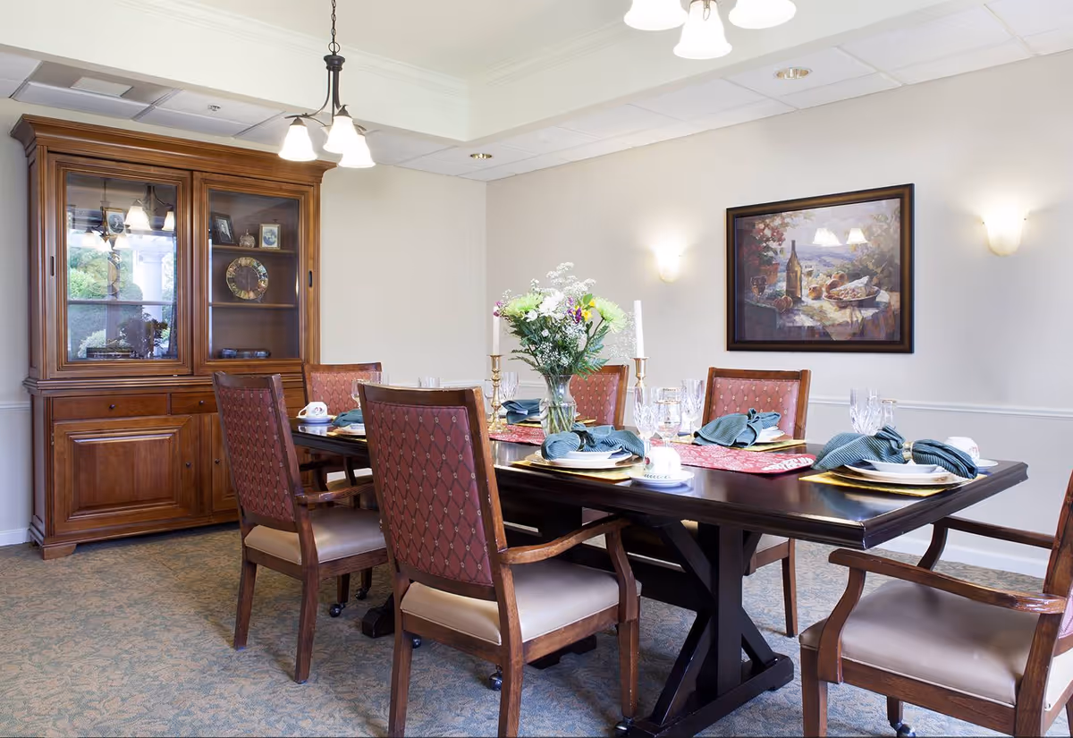 A formal dining room with a dark wooden table set for six people. The table has a floral centerpiece, candles, plates with folded napkins, and glassware. There are six upholstered chairs with wooden frames around the table. A wooden china cabinet with glass doors is against the wall, and a framed painting hangs on the adjacent wall. The room is softly lit by wall sconces and a ceiling light fixture.