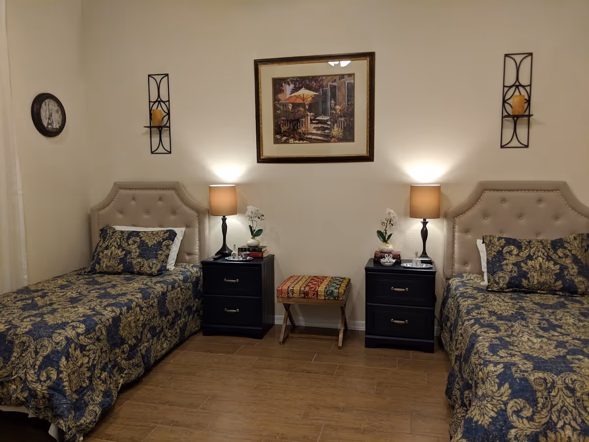 A bedroom with two twin beds featuring beige tufted headboards and dark blue and gold patterned bedding. Each bed has a black nightstand with two drawers, a table lamp, a small plant, and decorative items. A colorful cushioned bench is placed between the nightstands. The walls are light-colored, decorated with a framed painting above the bench and two wall-mounted candle holders on either side. A round wall clock is visible on the left wall.