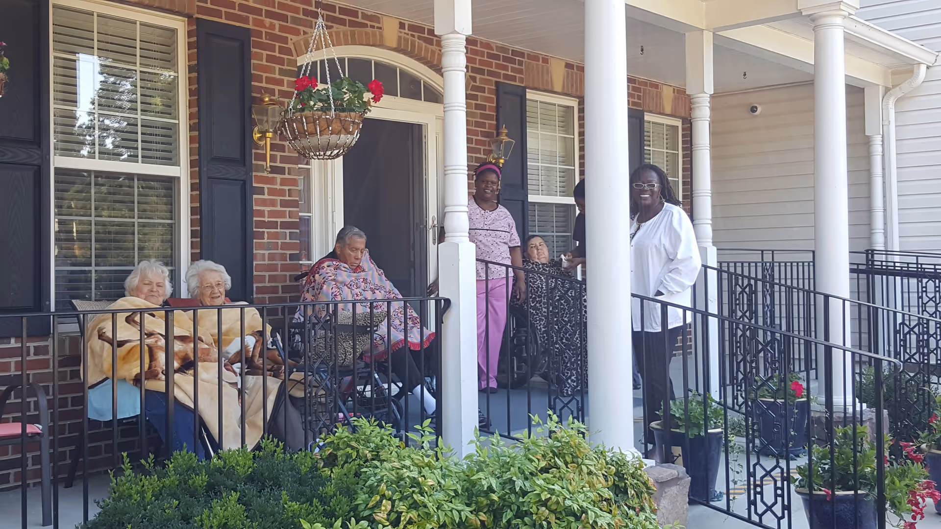A group of elderly people and caregivers gathered on a covered porch with brick walls and white columns. Some elderly individuals are seated in wheelchairs and chairs, wrapped in blankets, while caregivers stand nearby. There are potted plants and hanging flower baskets decorating the porch area.