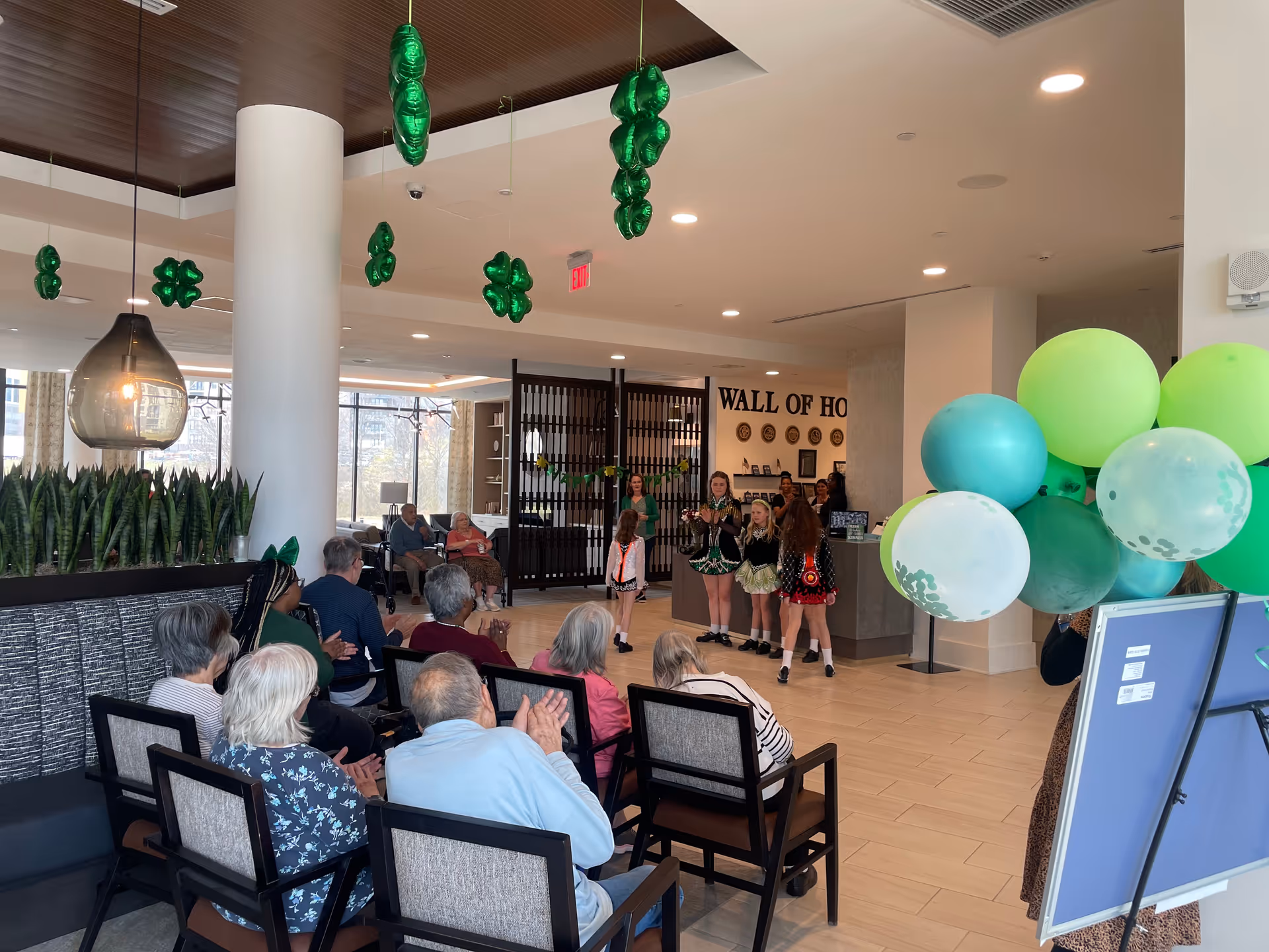 A group of elderly people seated and clapping in a common area of a senior living facility, watching a performance by young girls dressed in colorful costumes. The room is decorated with green balloons and hanging green shamrock decorations. There is a sign on the wall that reads 'WALL OF HO'.