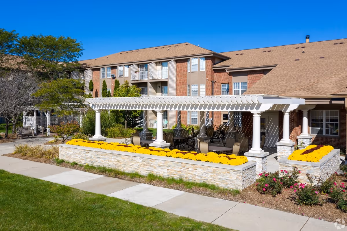 Outdoor seating area with a white pergola and several chairs and tables surrounded by flower beds with yellow and pink flowers, in front of a multi-story brick building under a clear blue sky.