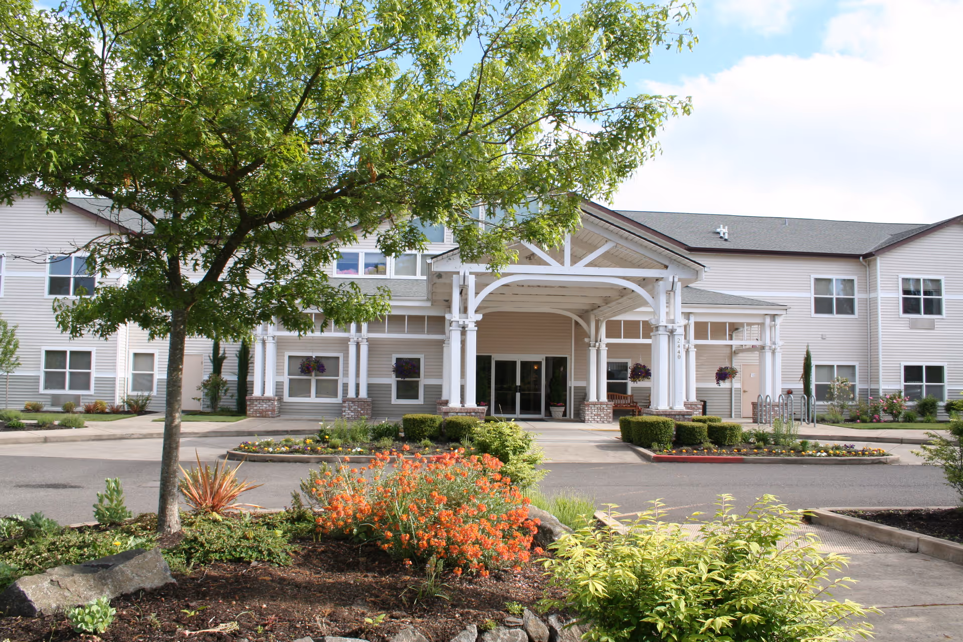 Front exterior view of a senior living facility with a covered entrance supported by white columns. The building is two stories tall with beige siding and multiple windows. There is a landscaped garden area with green shrubs, a tree, and orange flowers in the foreground under a partly cloudy sky.