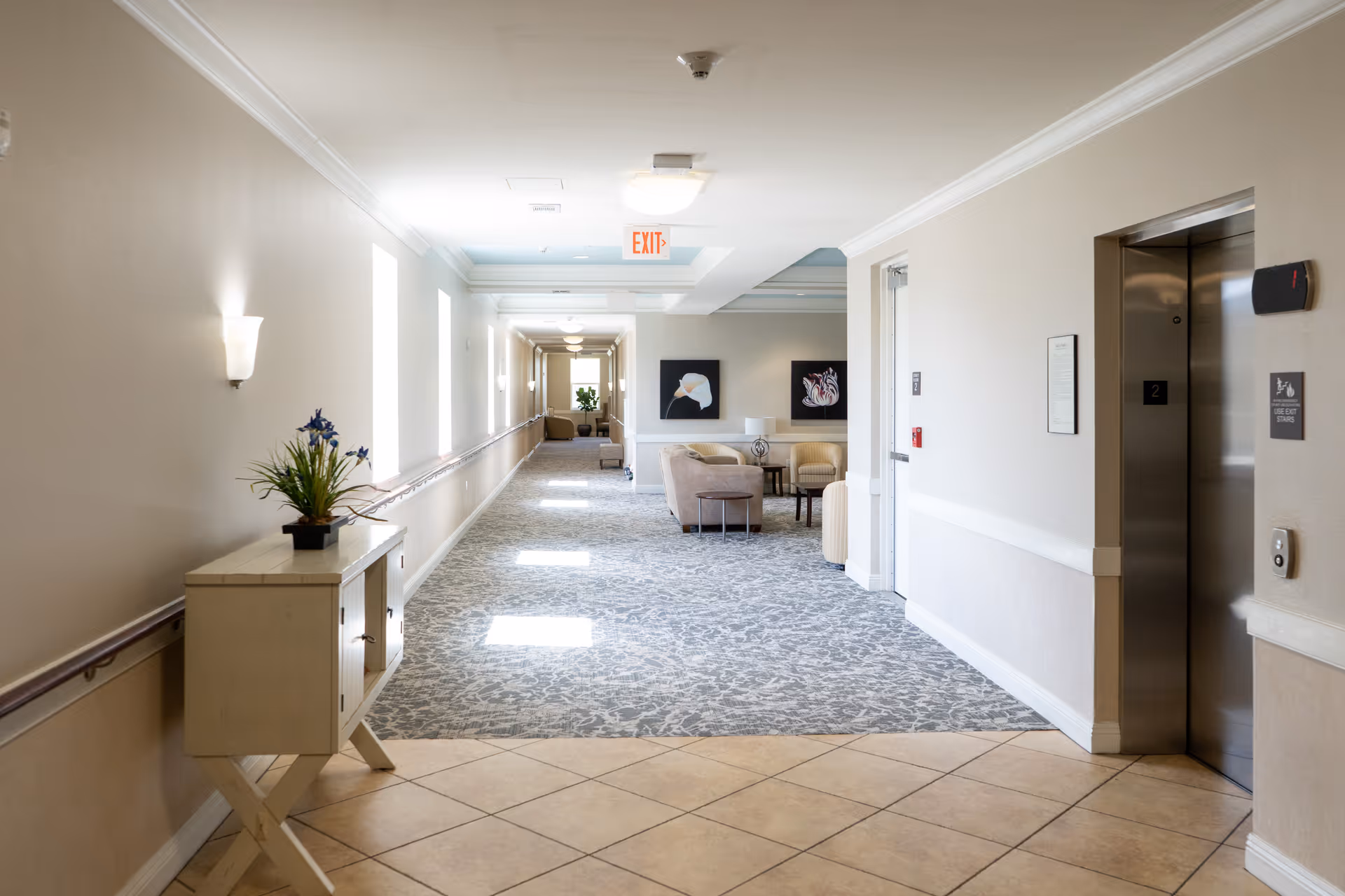 Bright, carpeted corridor and lounge area in a senior living facility with an elevator on the right.