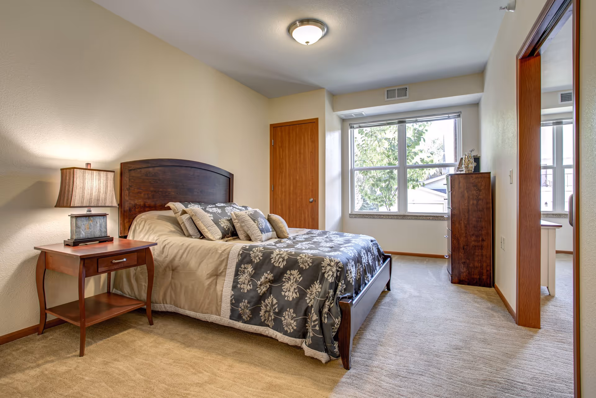 A neatly arranged bedroom with a wooden bed frame and a bed covered with a beige and floral patterned comforter. There are multiple pillows on the bed. A wooden nightstand with a lamp is placed beside the bed. The room has beige walls, a window with a view of trees outside, a wooden door, and a wooden dresser near the window. The floor is carpeted in a light beige color.