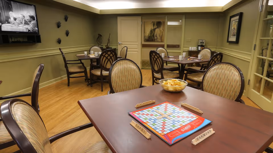 A cozy common room with several tables and cushioned chairs arranged around them. One table in the foreground has a Scrabble board game set up with wooden tile holders and a bowl of potato chips. In the background, a wall-mounted TV is playing a black and white show. The room has light green walls, wooden flooring, and framed artwork on the walls.