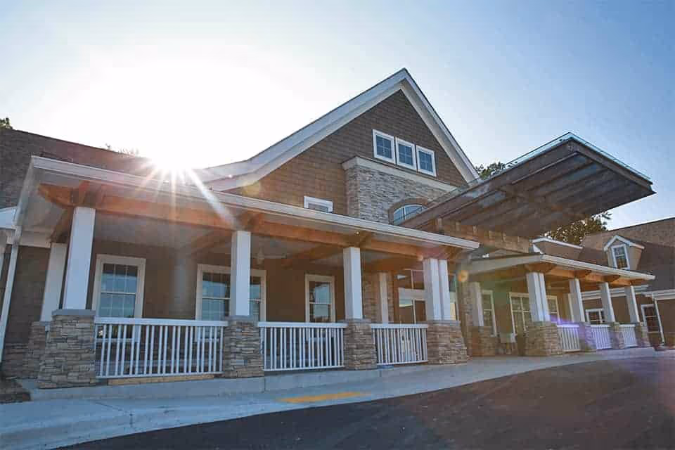 Exterior view of a senior living facility building with a covered entrance, stone and wood siding, white railings, and multiple windows under a clear blue sky with the sun shining brightly.