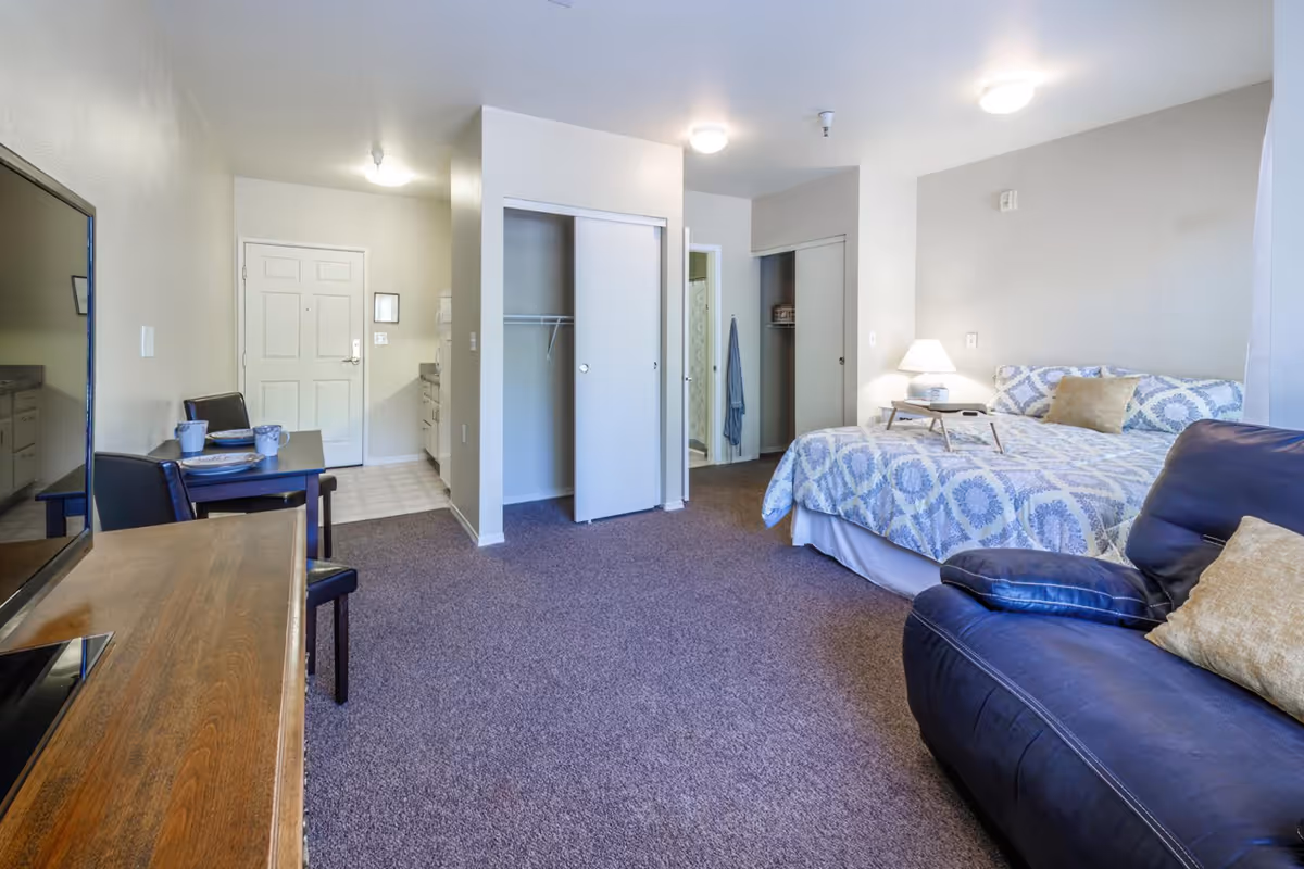 Interior view of a senior living facility room at Parkway Assisted Living featuring a bed with patterned bedding, a dark blue armchair with a beige pillow, a wooden TV stand with a flat-screen TV, a small dining table set for two, an open closet with sliding doors, and a doorway leading to a bathroom. The room has beige walls and carpeted flooring.