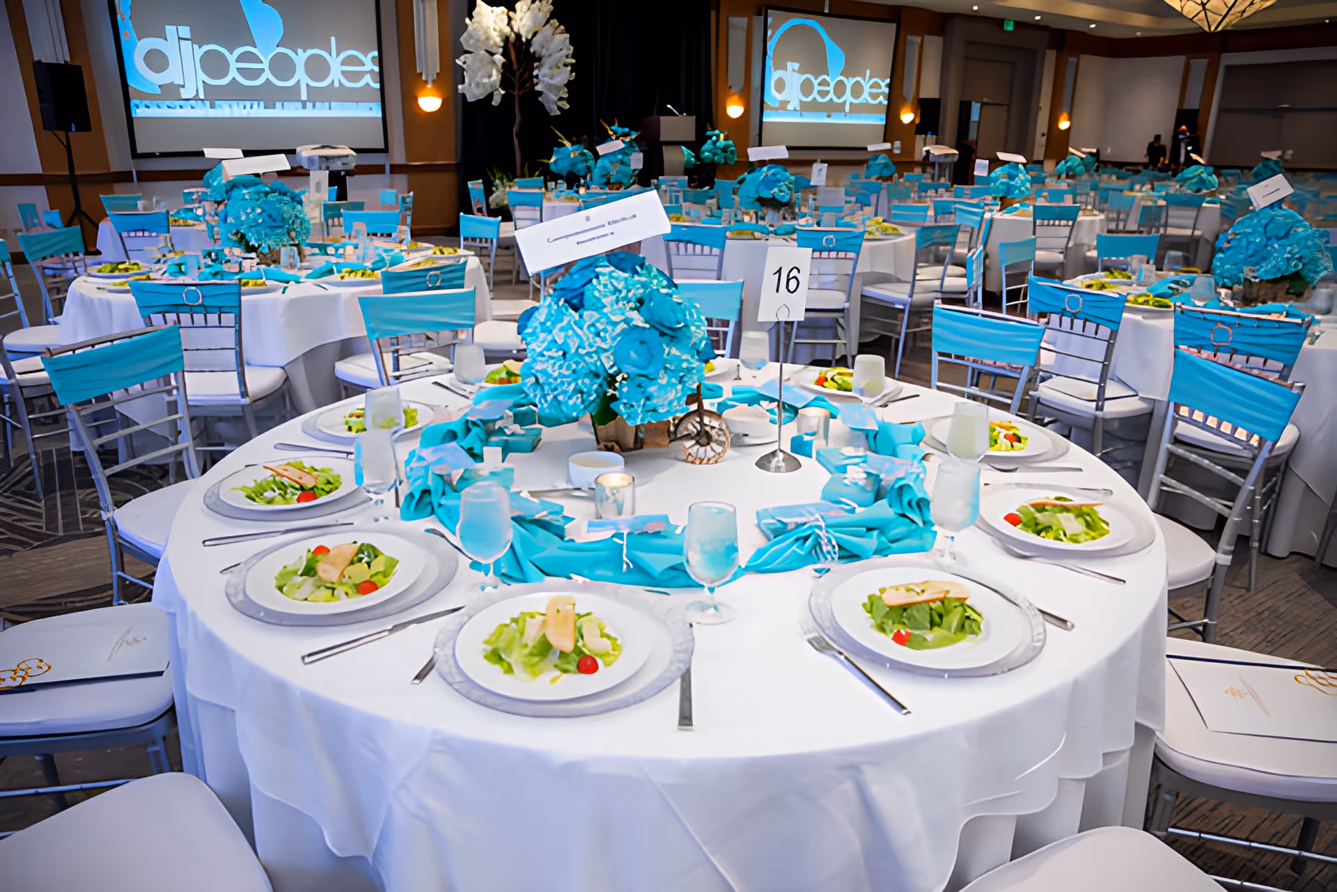 Round banquet tables set for a formal meal with turquoise floral centerpieces and salads on plates in a large event hall.