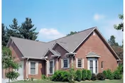 Exterior view of a single-story brick residential building with multiple windows, a gabled roof, and surrounded by green shrubs and trees under a blue sky.