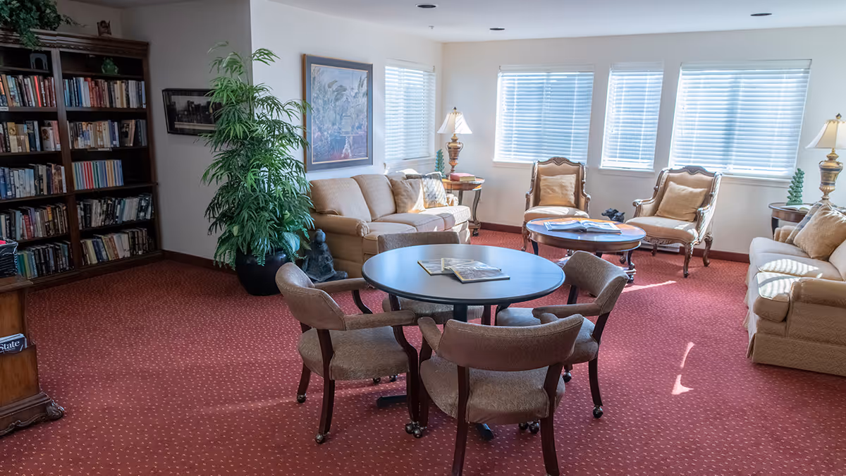 A cozy living room area with red carpet, beige sofas, and armchairs arranged around two round tables. One table has books on it, and there is a large potted plant near a bookshelf filled with books. The room is well-lit by natural light coming through multiple windows with white blinds.