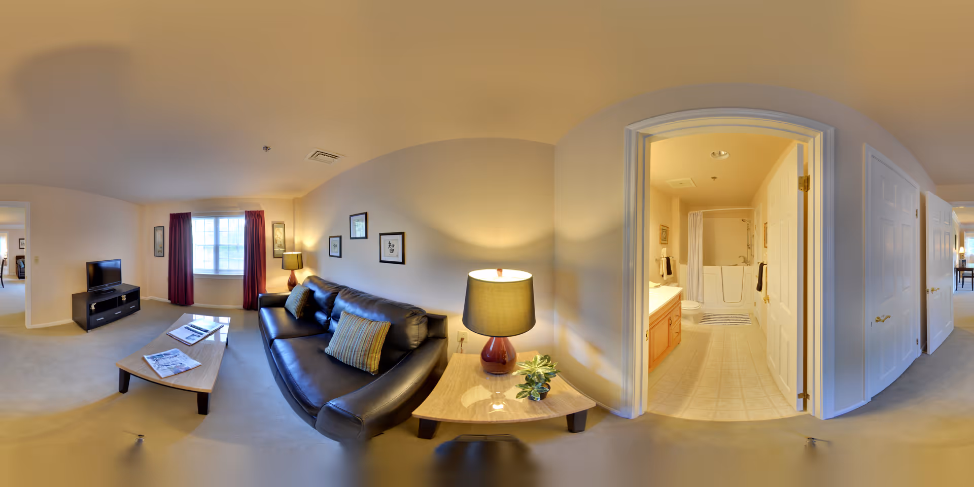 A spacious living area in a senior living facility featuring a black leather sofa with cushions, a wooden coffee table with newspapers, a side table with a lamp and small plant, a TV on a stand, and a window with red curtains. To the right, an open door reveals a bathroom with a walk-in bathtub, sink, and toilet. The walls are painted light beige, and the floor is carpeted in the living area with tile in the bathroom.