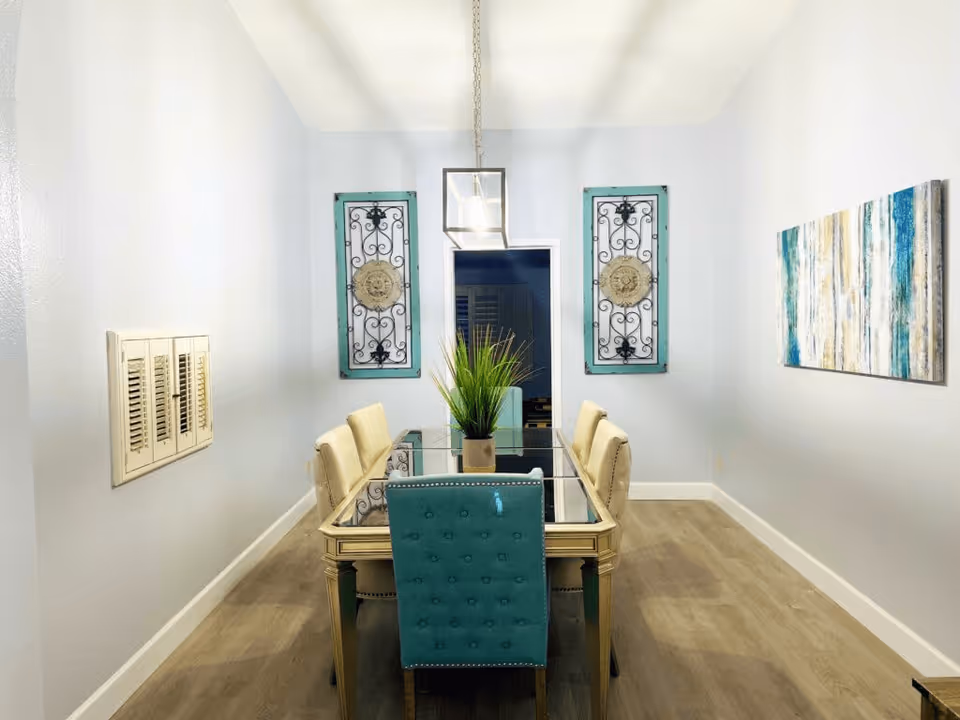 A dining room with a glass-top table surrounded by six chairs, five beige and one teal. The room has light-colored walls and wooden flooring. Two decorative wall hangings with intricate designs are on the far wall, flanking a doorway. A modern rectangular pendant light hangs above the table, and a colorful abstract painting is on the right wall.