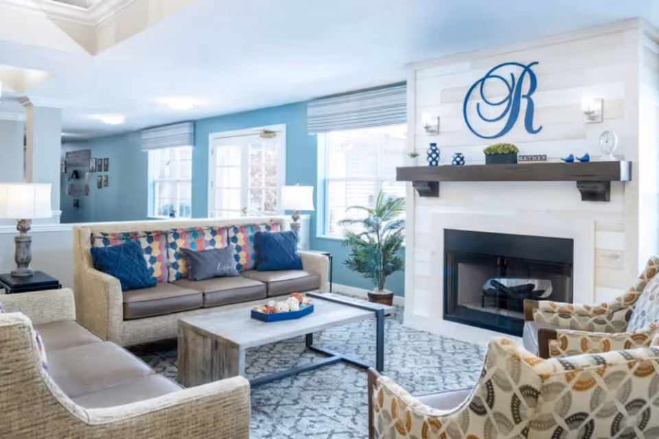 A bright and cozy living room with light blue walls and large windows letting in natural light. The room features a beige sofa with colorful cushions, two patterned armchairs, a wooden coffee table, and a fireplace with a wooden mantle decorated with blue vases, a plant, and a large decorative letter 'R' above it. There are also two table lamps on side tables and a patterned rug covering the floor.