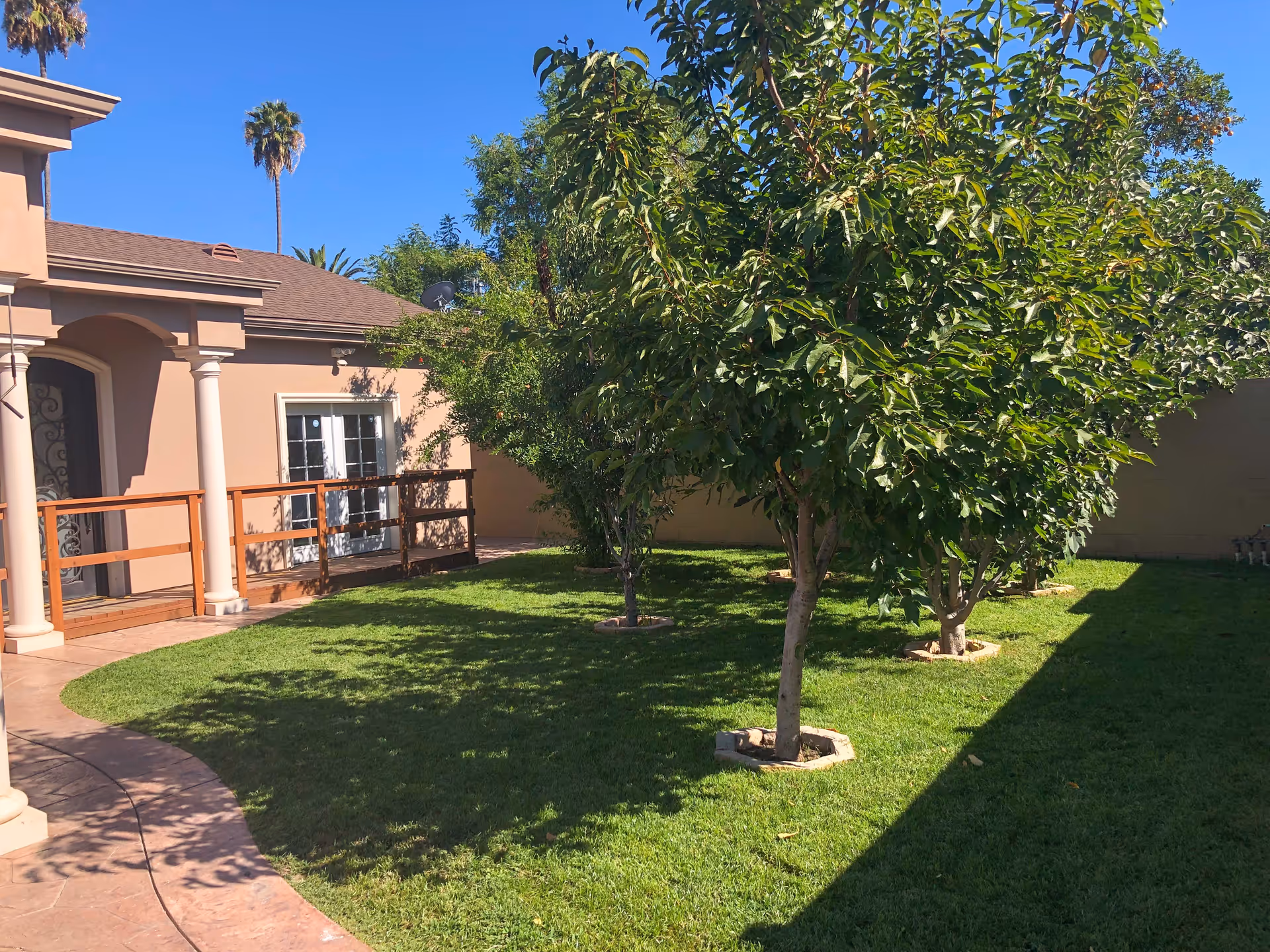 A sunny outdoor garden area at Sweet Home Senior Living with a well-maintained lawn, several small trees planted in circular stone borders, and a beige building with a wooden railing and white columns in the background under a clear blue sky.