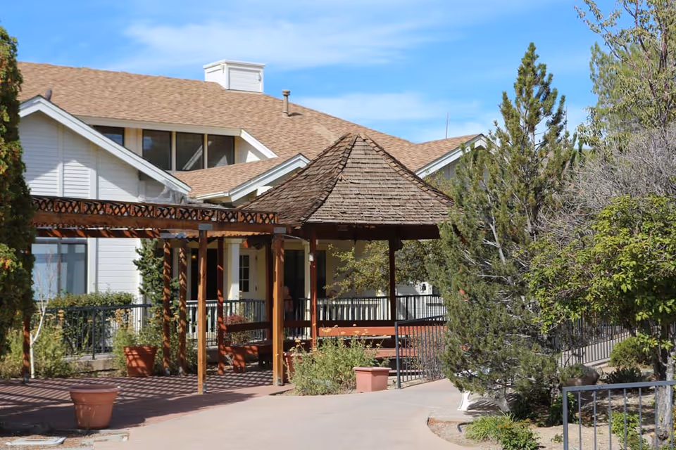 A courtyard with a wooden gazebo and pergola in front of a multi-story assisted living building, surrounded by trees and potted plants.