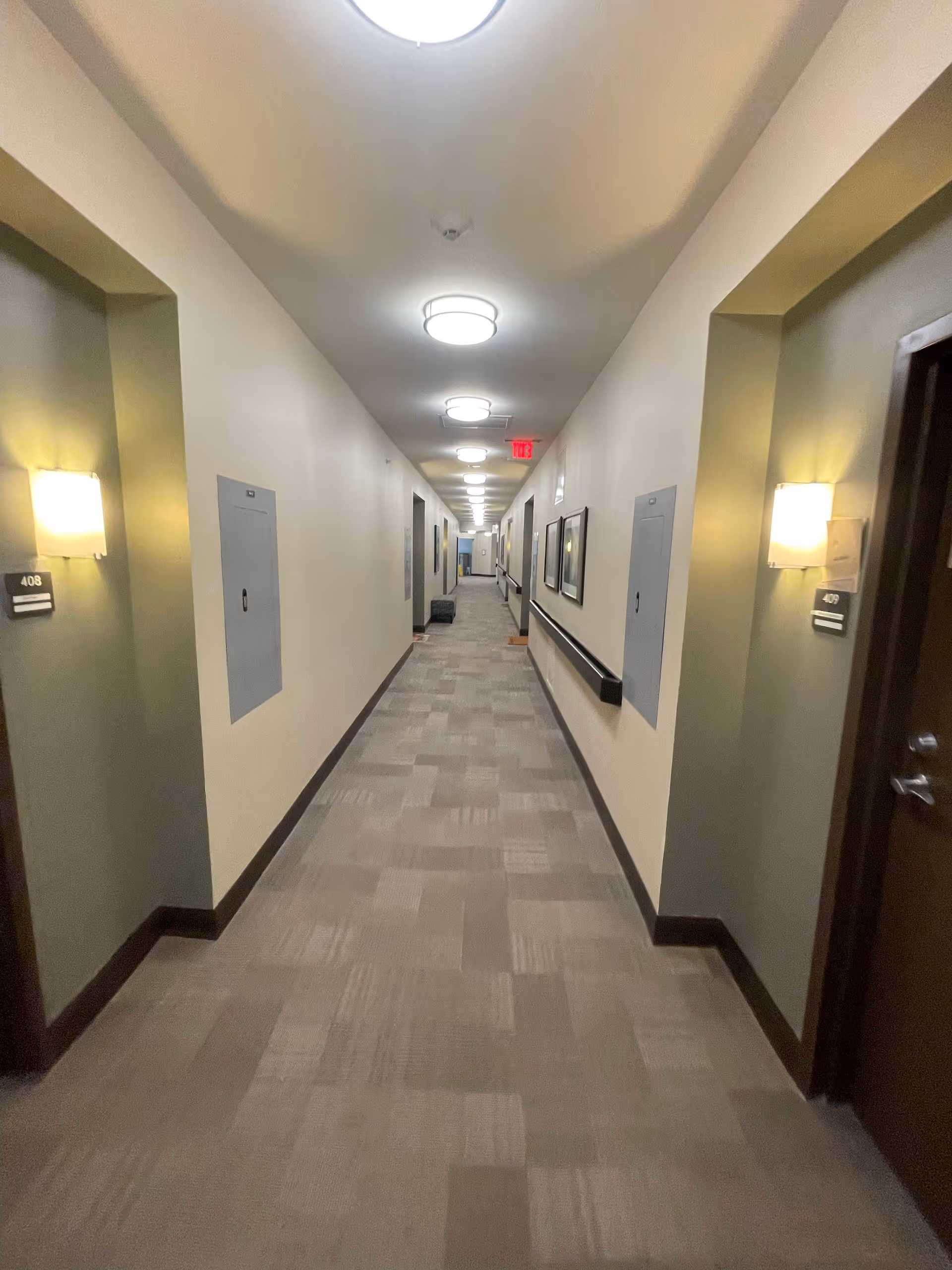 A long, well-lit hallway in a senior living facility with beige walls and carpeted floor. Doors to rooms 408 and 409 are visible on either side near the foreground, with wall-mounted lights above the room number plaques. The hallway has ceiling lights evenly spaced along the length and handrails on the right side wall. Some framed pictures are hung on the right wall further down the corridor.