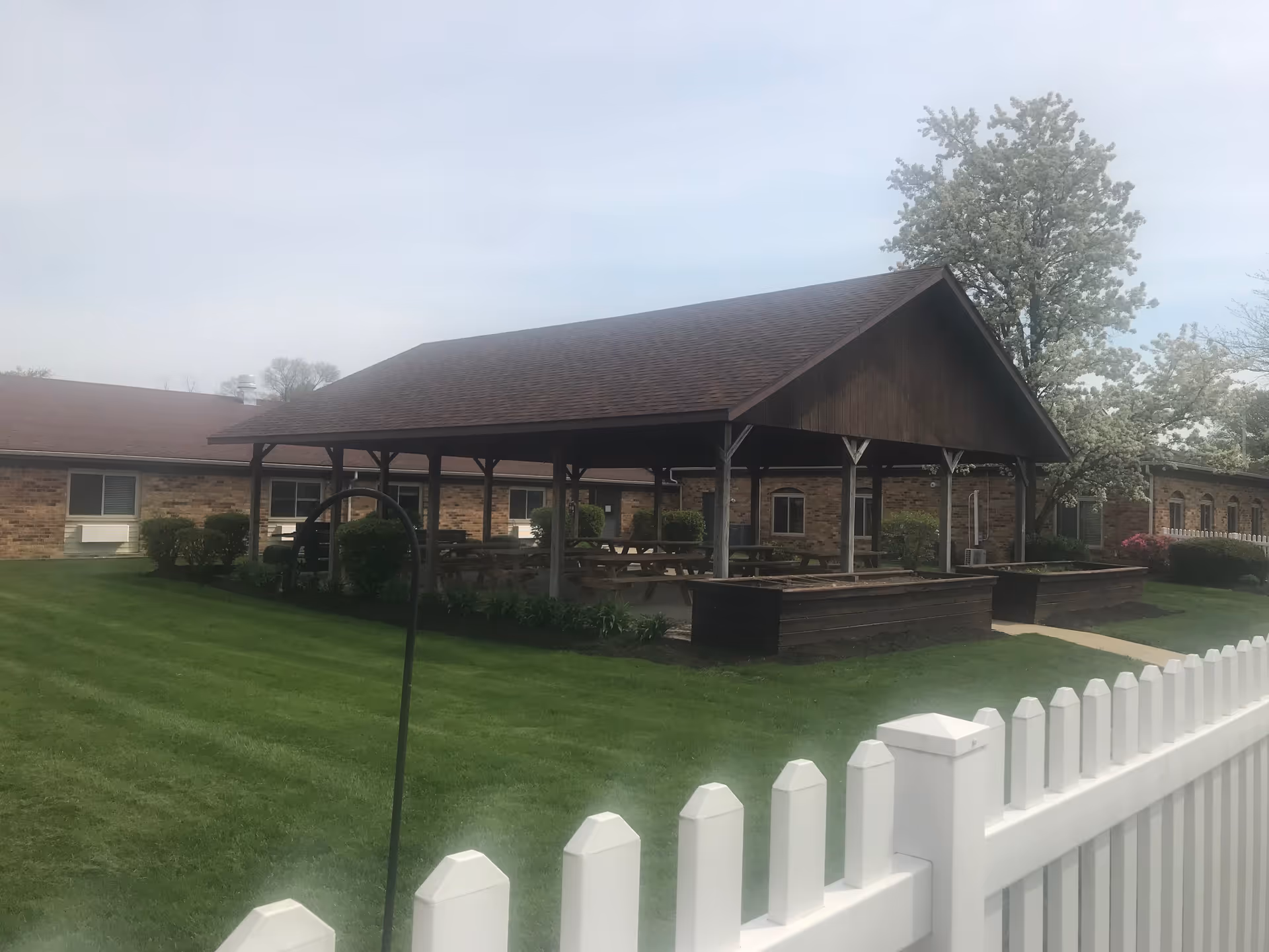 Outdoor covered pavilion with picnic tables surrounded by well-maintained green grass and landscaping, adjacent to a brick building with windows. A white picket fence is visible in the foreground, and a tree with white blossoms is in the background.