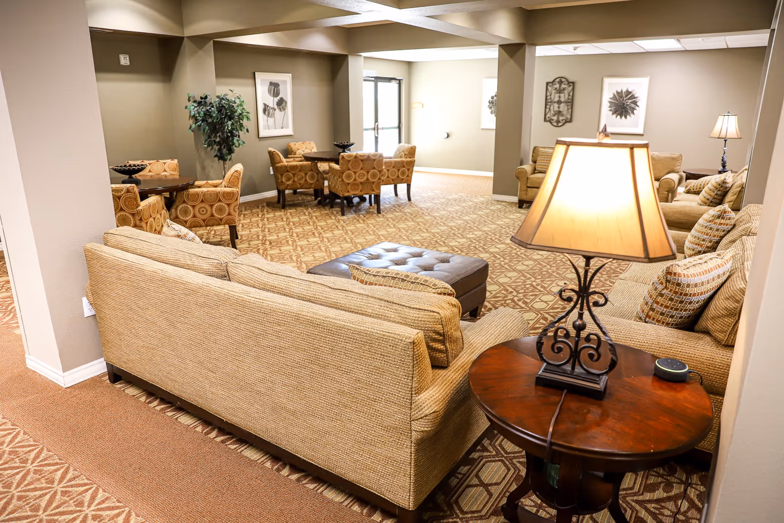 A spacious living room area in a senior living facility with beige sofas, patterned armchairs, a wooden side table with a lamp, and framed artwork on the walls. The room has patterned carpet flooring and soft lighting, creating a warm and inviting atmosphere.
