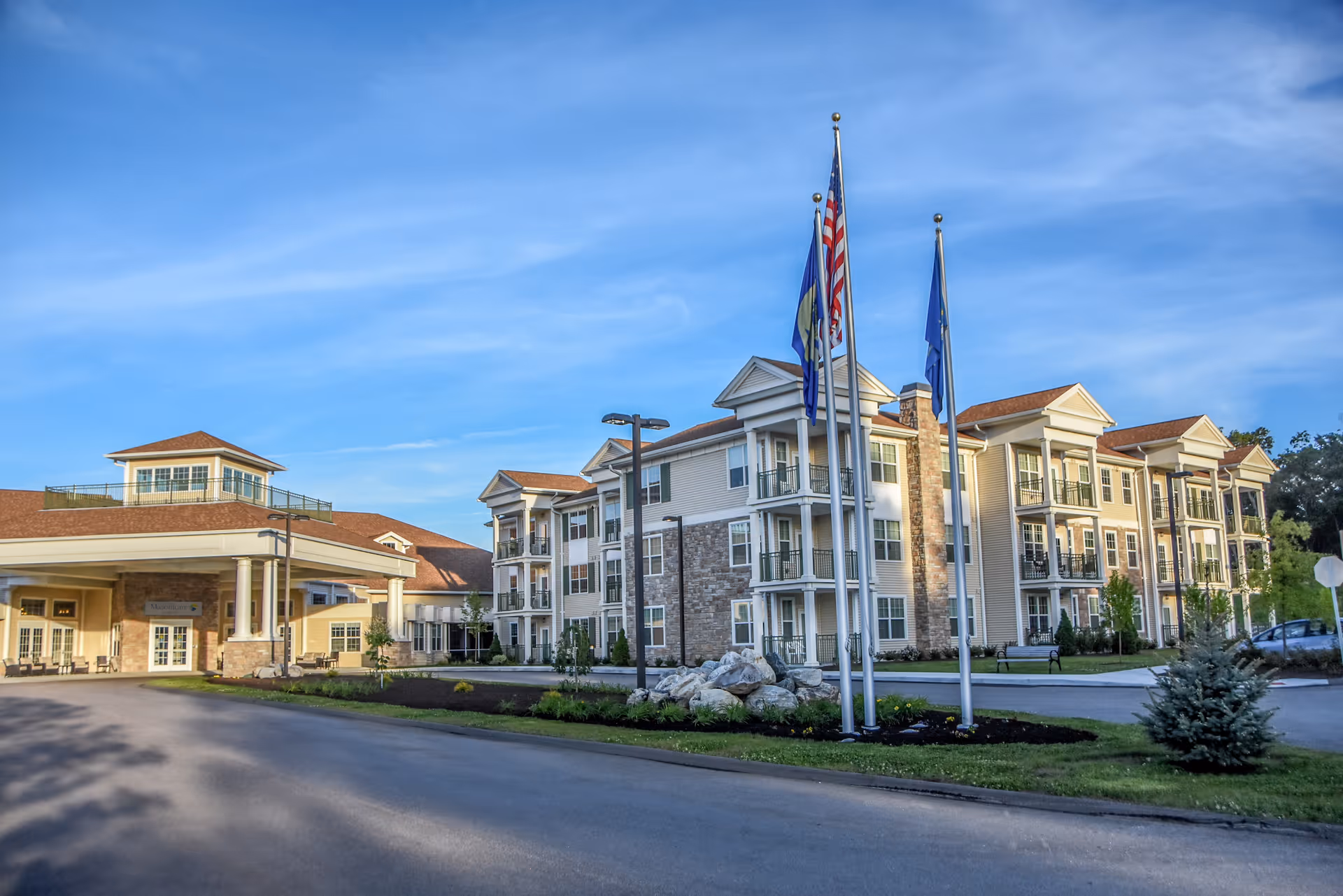 Exterior view of a senior living facility named Masonicare At Mystic, showing a large multi-story building with balconies, a covered entrance, three flagpoles with flags, and a clear blue sky.