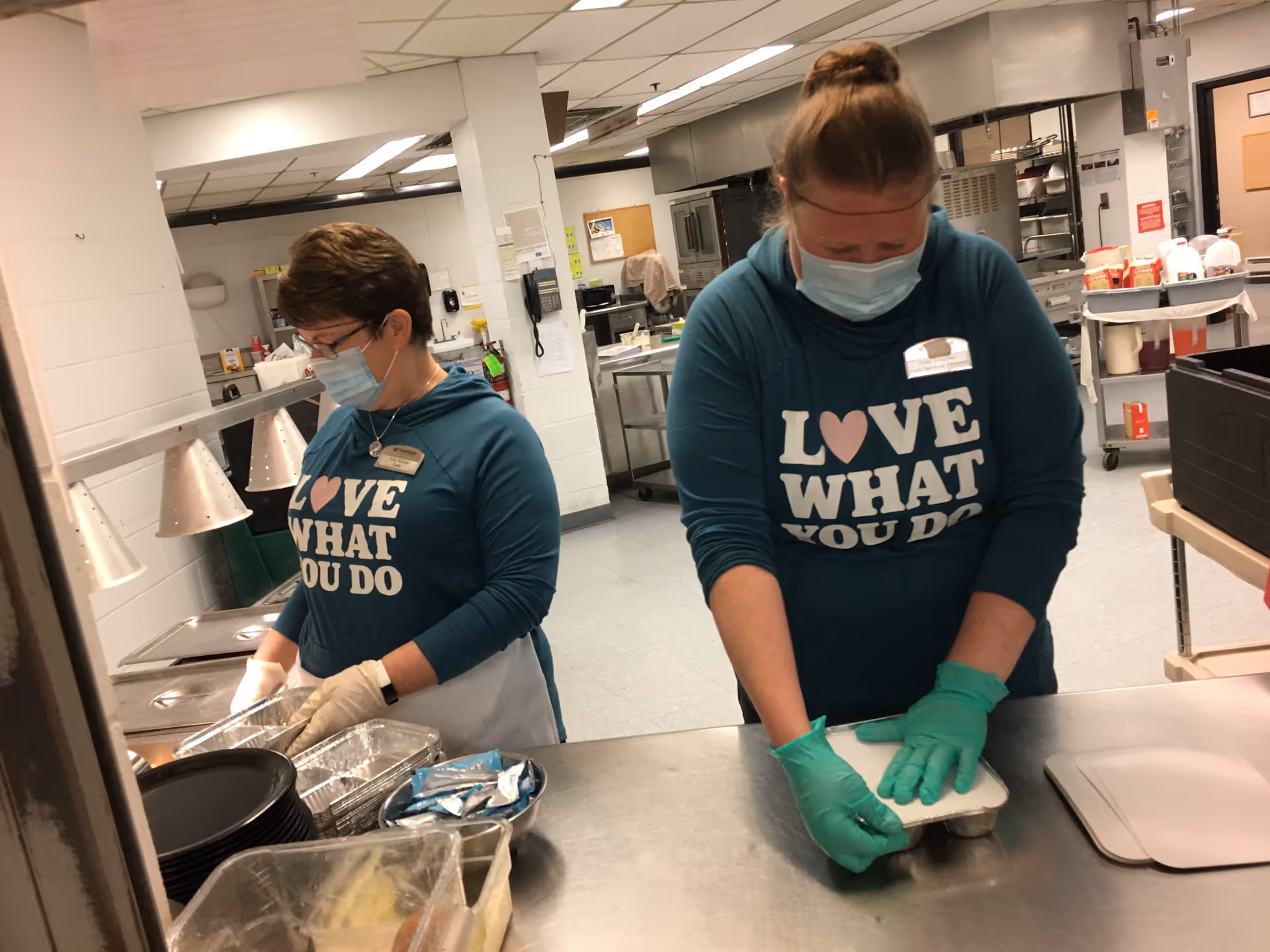Two women wearing teal hoodies with the phrase 'LOVE WHAT YOU DO' and face masks are working in a commercial kitchen. One woman is handling aluminum trays while the other is sealing a food container. The kitchen has stainless steel counters and various kitchen equipment in the background.
