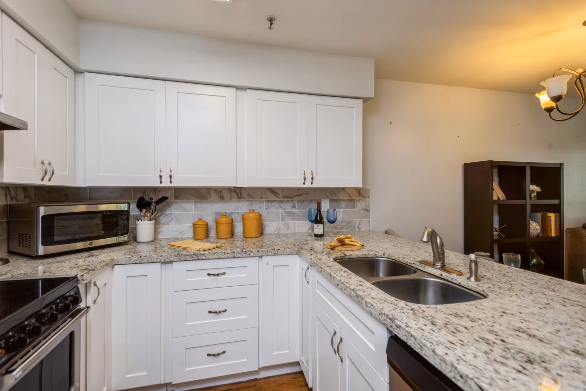 Modern kitchen with white cabinets, granite countertops, a double sink, a microwave, and a stove. There are three yellow canisters, a bottle of wine with two blue wine glasses, and some kitchen utensils on the counter. In the background, there is a dark brown shelving unit with books and decorative items.