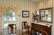 A dining room with a glass-top table and wooden chairs. The room has striped wallpaper, a window with floral curtains, a large mirror above a wooden sideboard, and decorative items including a vase with flowers and two lamps.
