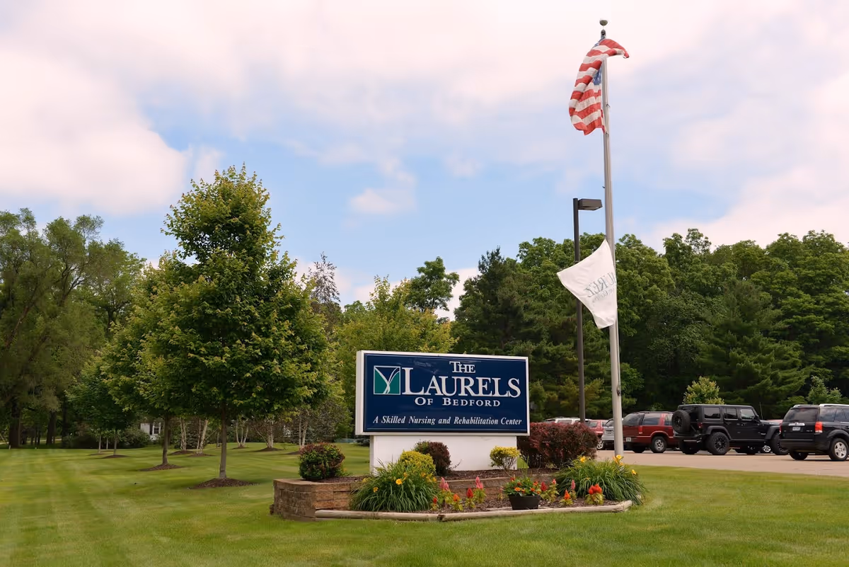 Outdoor view of The Laurels of Bedford skilled nursing and rehabilitation center sign surrounded by green grass, trees, and flowers with an American flag and another flag on a flagpole nearby. Several parked cars are visible in the background under a partly cloudy sky.