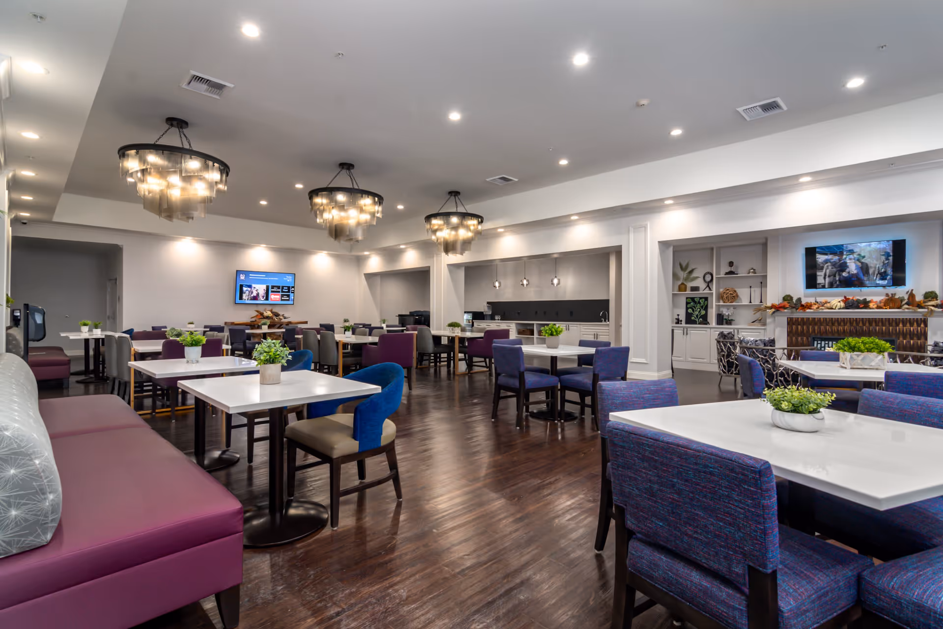 A spacious dining room with multiple tables and chairs arranged neatly. The room features modern chandeliers hanging from the ceiling, a dark wooden floor, and a mix of purple and blue upholstered chairs. There are small green plants placed on each table, a wall-mounted TV, and decorative shelves with plants and ornaments in the background.