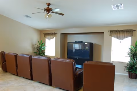 A living room with a row of six brown leather recliner chairs facing a large flat-screen TV mounted in a recessed wall. The room has beige walls, two windows with valance curtains, two potted plants on either side of the TV, and a ceiling fan with lights.