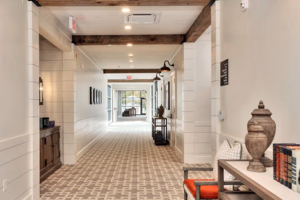 A well-lit hallway in a senior living facility with patterned carpet, white shiplap walls, and wooden ceiling beams. The hallway features decorative items such as vases, books, and framed pictures on the walls. There are benches with cushions along the right side and a wooden cabinet on the left. At the end of the hallway, large windows allow natural light to enter, revealing a seating area with chairs.