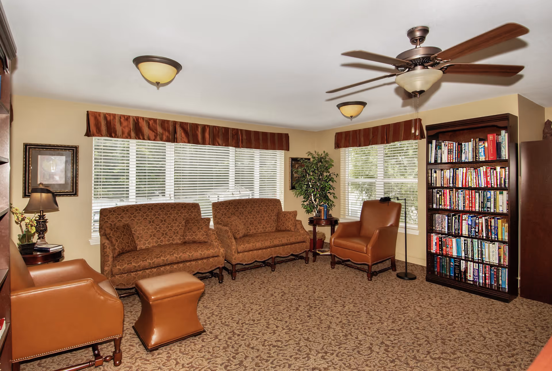 A cozy living room with two patterned brown sofas, two brown leather armchairs, a matching ottoman, a wooden bookshelf filled with books, a small side table with a lamp and plant, and large windows with blinds and red valances. The room has beige walls, a patterned carpet, and ceiling lights including a ceiling fan with a light.